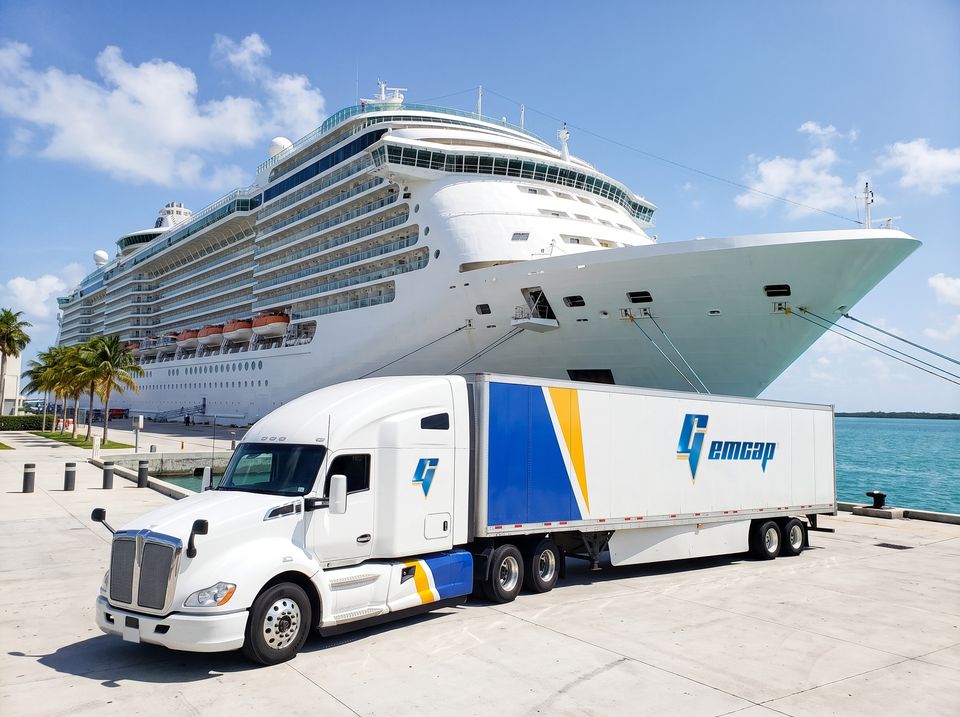 A white GEMCAP tractor-trailer parked on the pier at PortMiami beside a docked cruise ship