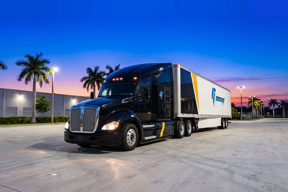 A black GEMCAP tractor-trailer with its headlights on in the Miami truck yard at blue hour
